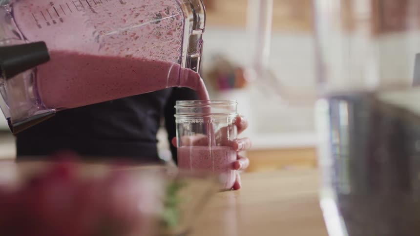 A woman pours a berry smoothie from a blender into a glass jar