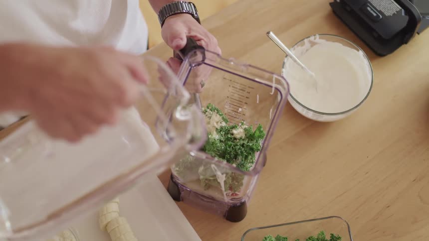 A person pours a water from a pitcher into a blender filled with vegetables