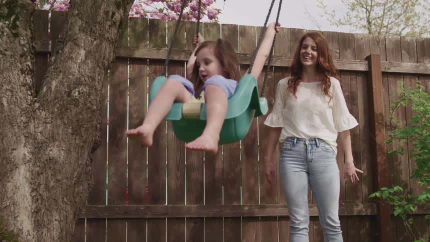 A woman pushing a girl on a swing with a fence and trees in the background