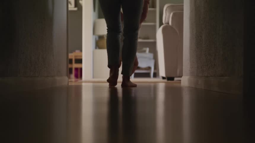 A mother slowly walks down a hallway helping her child who is just learning to walk