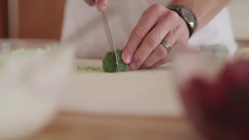 Man slices broccoli florets on pale chopping board at wooden table