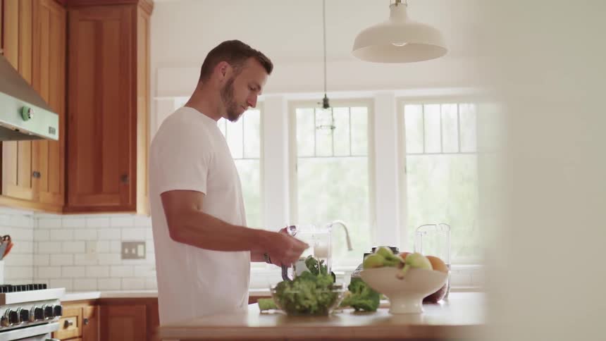 A man in a kitchen putting powder in a blender with vegetables