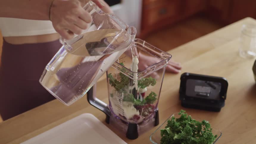 A young woman at home pours water in to a blender as she makes a smoothie