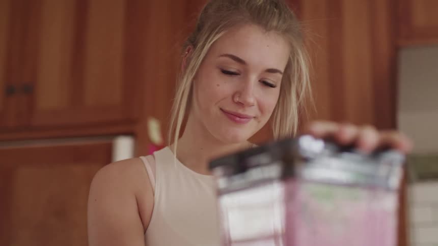 Blonde woman with a white short sleeve t shirt blends up a smoothie with a blender in the kitchen
