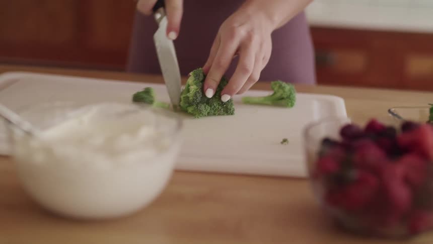 A woman chops broccoli on a cutting board as a bowl of whipped cream and a bowl of fruit sit nearby