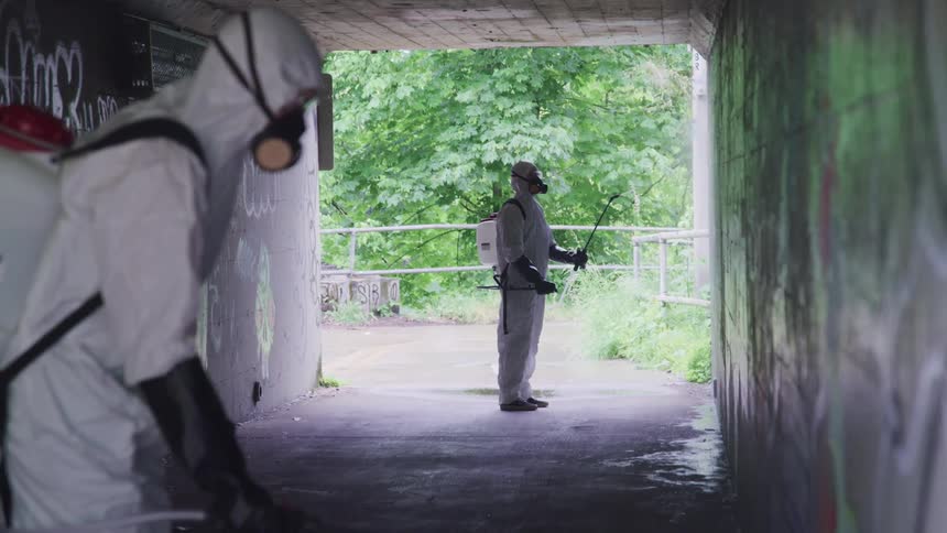 Two people stand in graffiti covered concrete tunnel wearing protective gear as they spray the walls