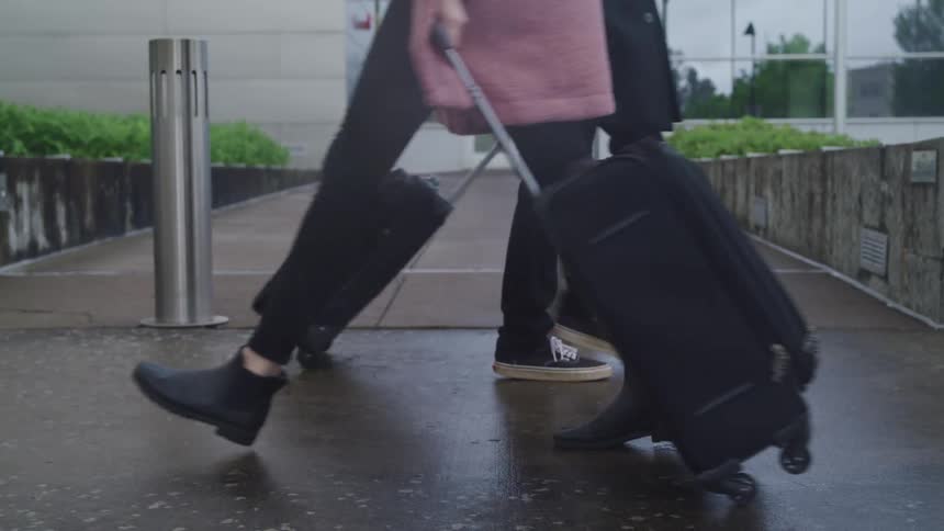 A pair of pedestrian pulling luggage behind them pass each other on a rain soaked road