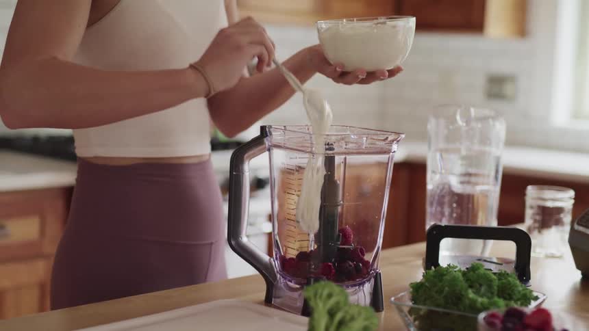 A woman pours cream into a blender filled with raspberries