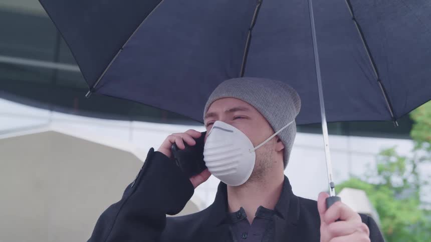 Man wearing face mask talks on a smartphone while holding an umbrella