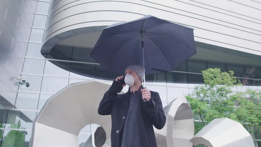 Man with a grey beanie a white face mask and a black overcoat holding a black umbrella stands and talks on the smartphone in front of an office building