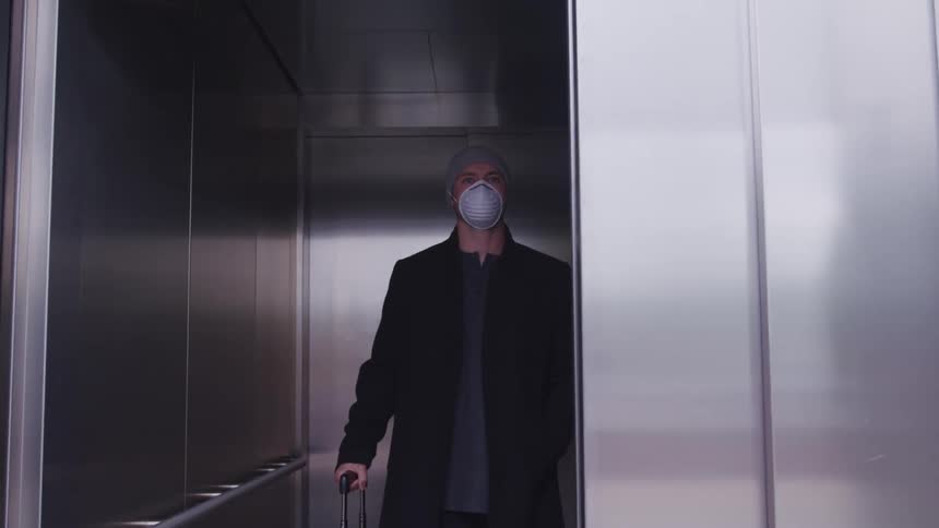 A man wearing a dust mask stands with his luggage on an elevator and looks out of the elevator towards the camera as the door closes