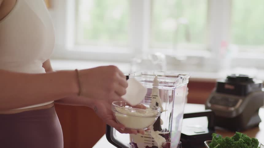 A woman wearing fitness apparel pours powder from a bowl into a blender using a plastic measuring cup