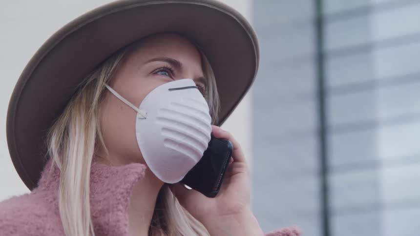 Blonde woman with pink overcoat a hat and a white face mask talks on a smart phone