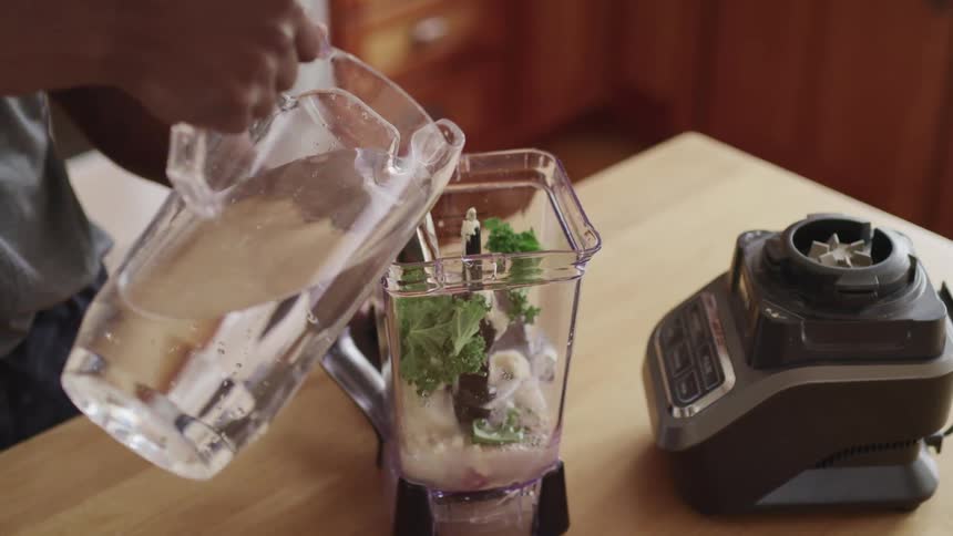 A man pours water from a clear pitcher into a blender filled with vegetables