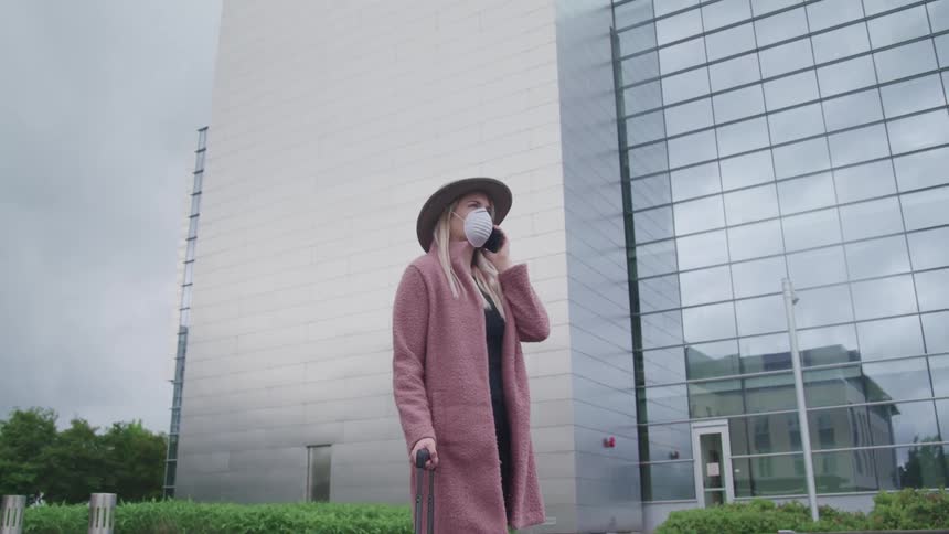 A woman stands outside of a tall building wearing a sun hat medical mask and hold a cell phone and cane