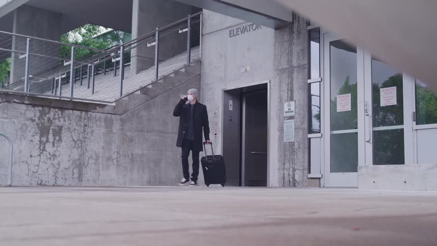 A man holding a smartphone to his ear and pulling luggage behind him exists an elevator and walks out of frame