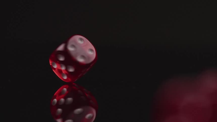 Red die with white spots spins on end on dark reflective surface