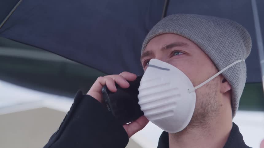 Man with a grey beanie and a white face mask holds a black umbrella and talks on a smart phone