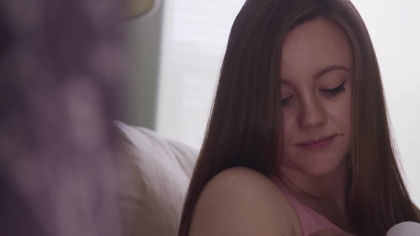 Mother with straight brown hair sitting in a white armchair holds her baby and looks at it affectionately in a room at home
