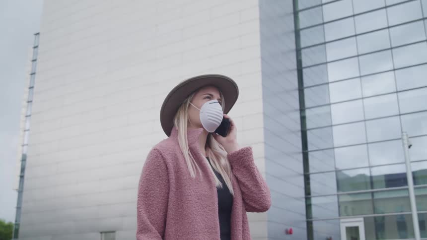 Woman wearing a medical mask holds a cell phone to her ear while standing near a tall building in a city