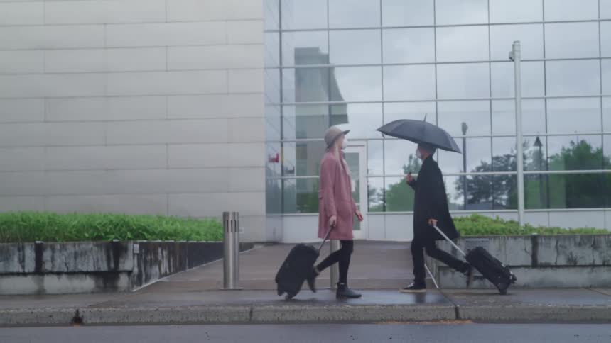 Woman with a face mask a pink coat and a hat and a man with a face mask and black umbrella walk on the pavement
