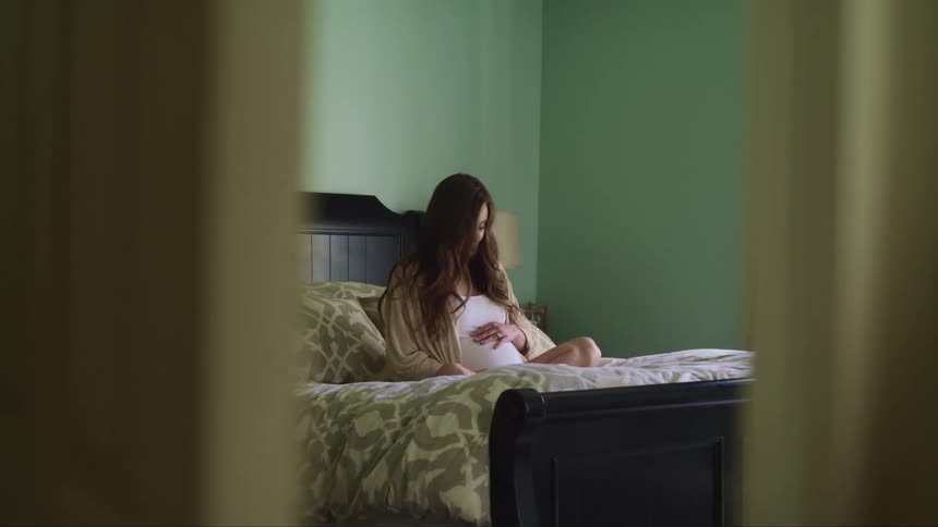 Pregnant woman with long brown hair sits on a bed and holds her belly in the bedroom at home
