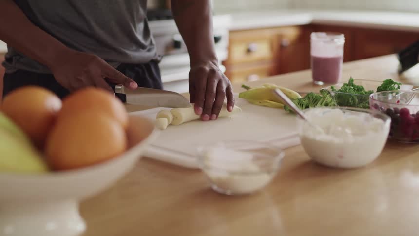 A man cuts up a banana and is surrounded by ingredients to make a smoothie