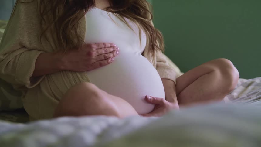 Pregnant woman wearing white t shirt sits in bed touching her belly in the bedroom