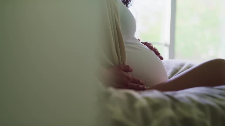 Pregnant woman with a white shirt and brown hair sits on a bed and holds her belly in bedroom