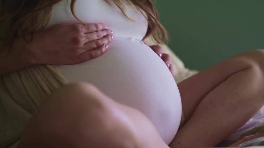 Pregnant woman with brown hair sits on a bed and strokes her belly