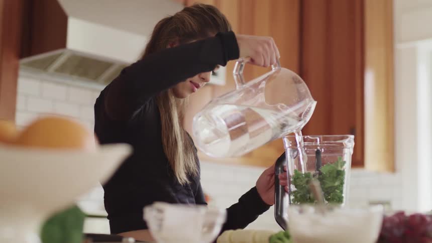 Woman pours water from a pitcher into a blender jar that contains green food