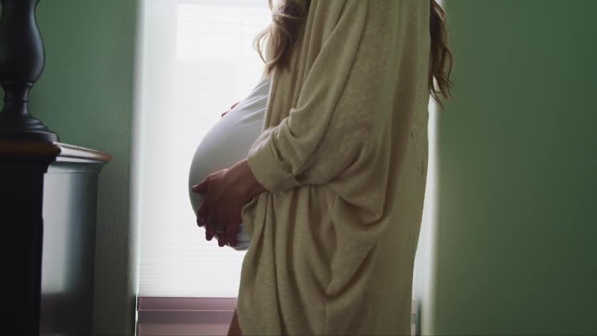 Pregnant woman with long brown hair stands and holds her belly in a room