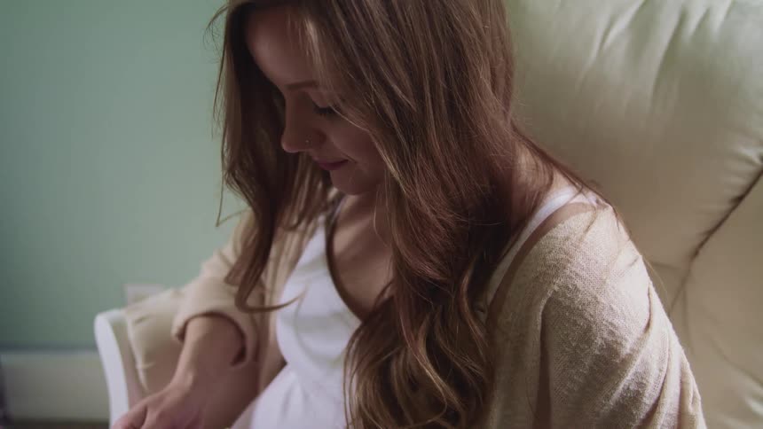 Pregnant woman with long brown hair sits on a white sofa at home