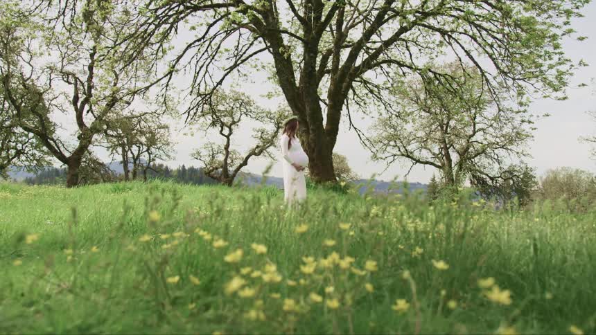 Pregnant woman with a white dress and a flower crown standing on grass under a tree holds her belly