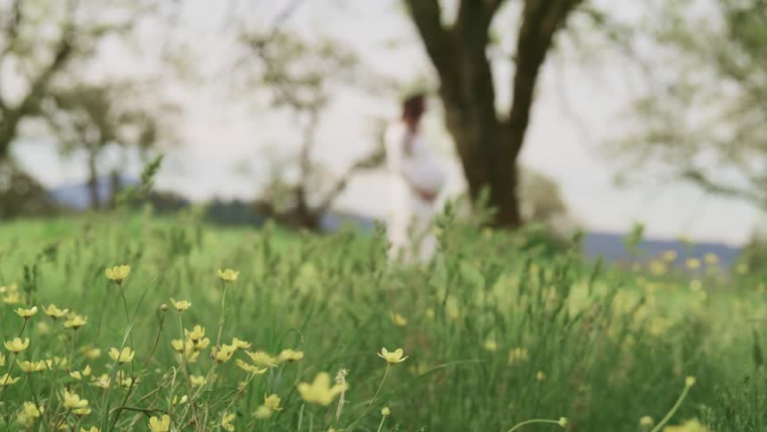 Pregnant woman with a white dress standing on grass under a tree holds her belly