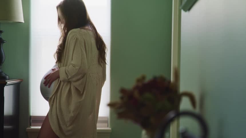 Young pregnant woman stands near a window in a room at home