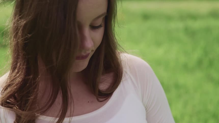 Woman with brown hair and a white long sleeve t shirt stands on the grass and looks down