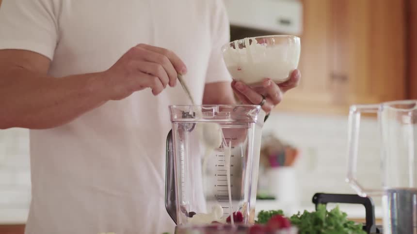Man with a white t shirt puts pale yellow cream into a blender to make a smoothie in the kitchen at home