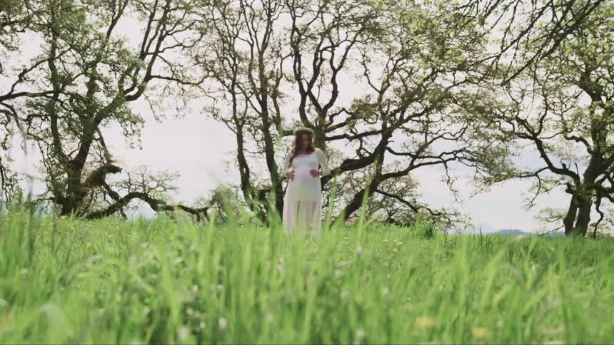 Pregnant woman with a white dress flower crown and long brown curly hair stands on the grass and strokes her belly in the forest