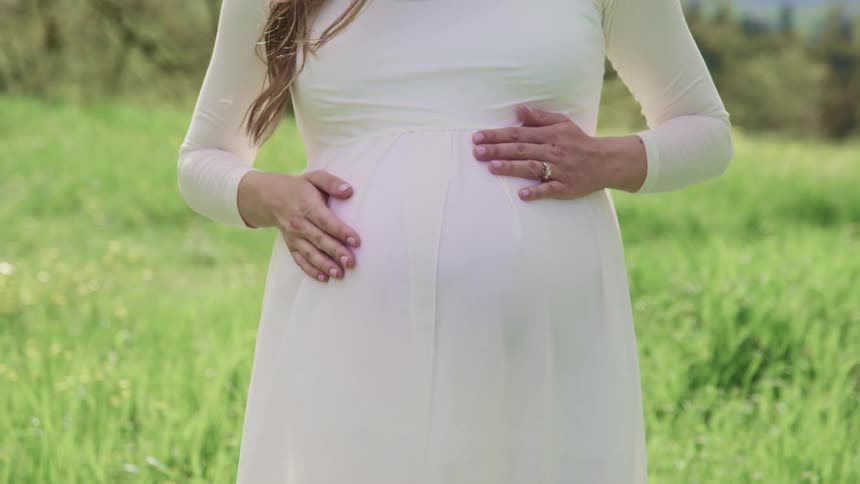 A pregnant woman stands in a field in a white dress and cradles her stomach