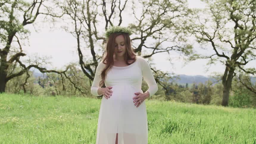 Pregnant woman with a white dress flower crown and long brown curly hair stands on the grass and strokes her belly in the forest