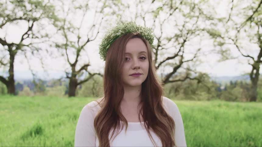 Woman dressed in white and wearing garland stands in field near trees