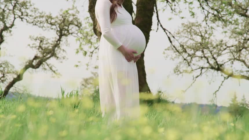 A heavily pregnant woman stands in a long white dress in an open field and cradles her stomach