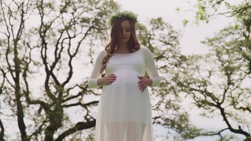 Pregnant woman with a white dress flower crown and long brown curly hair strokes her belly