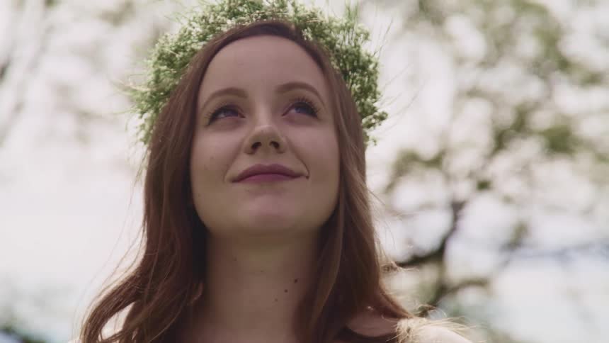 A woman wearing a flower crown outdoors looks up toward the sky and smiles