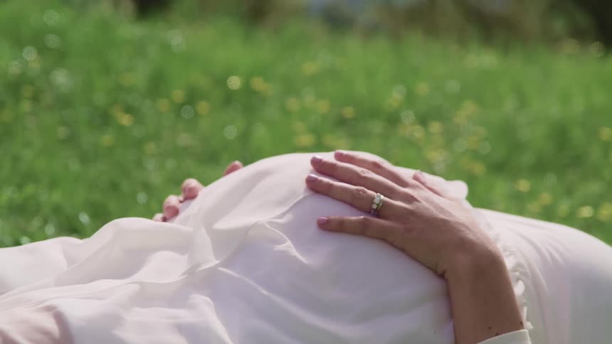 Pregnant woman in a white dress lays down on a grass field rubbing her belly