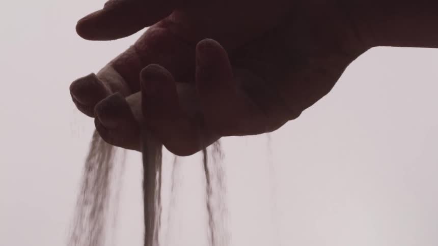 Sand runs through finger of hand falling against pale background
