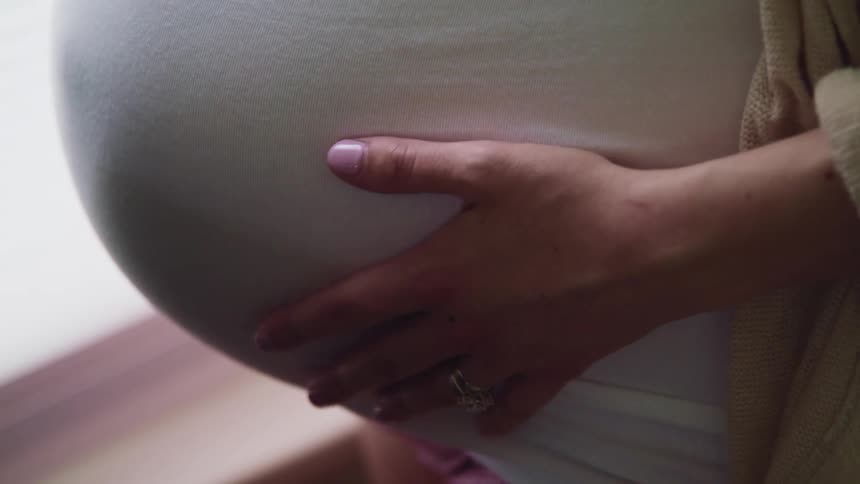 A pregnant woman stands in front of a window and embraces her pregnant belly