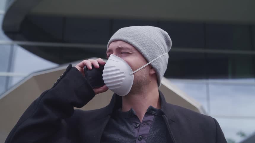 A man uses a smartphone while standing outside of a building wearing a dust mask