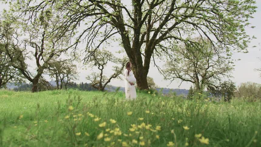Pregnant woman with a white dress flower crown and long brown curly hair holds her belly